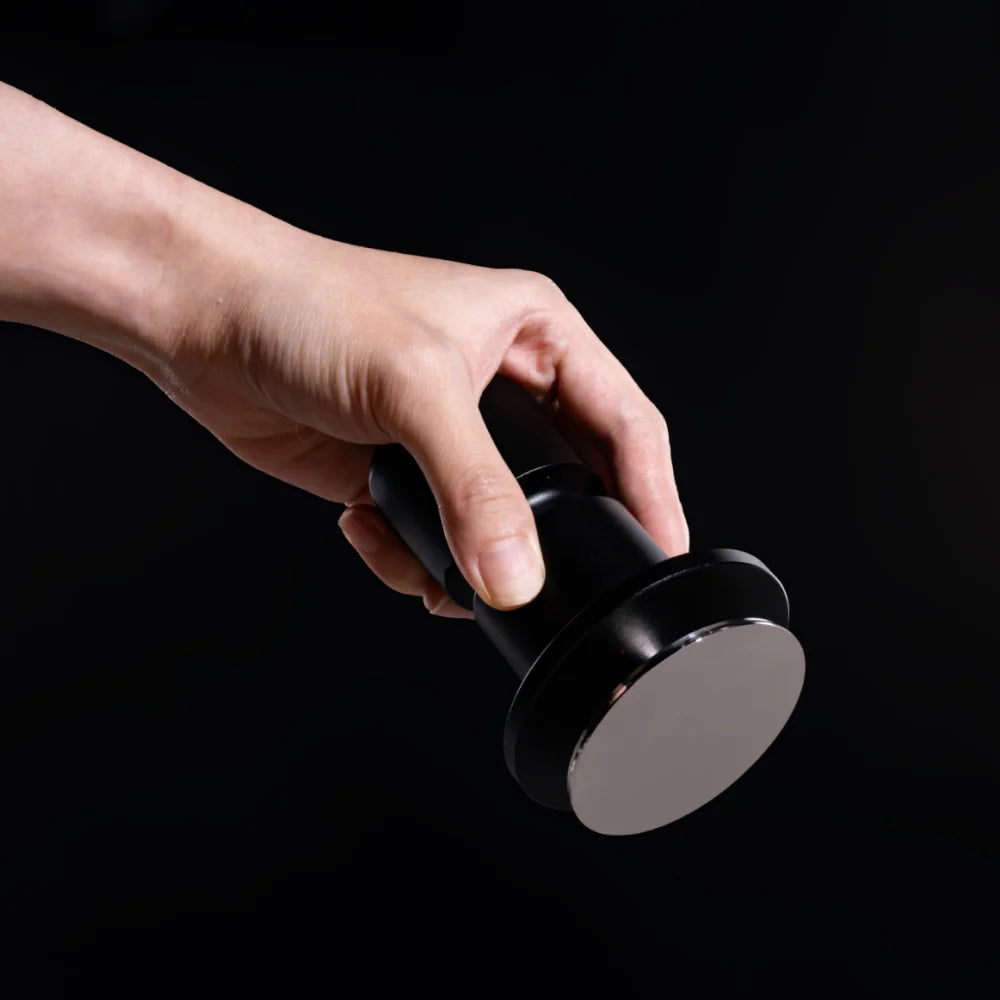A hand holding the Impulse Impact Tamper tilted to show its polished stainless steel base against a black background