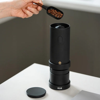 Coffee beans being scooped into the open top of the TIMEMORE x MILLAB E01 on its stand, lid set aside
