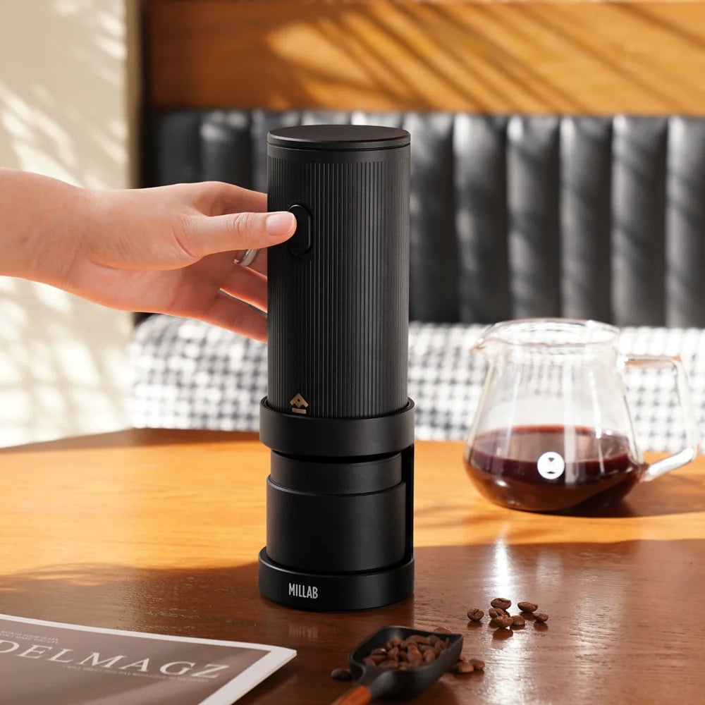 Hand pressing the power button of the TIMEMORE x MILLAB E01 on its stand, glass server of filter coffee behind