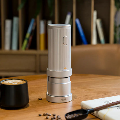 TIMEMORE x MILLAB E01 on its stand on a wooden table in front of a bookshelf, beside a cup of coffee