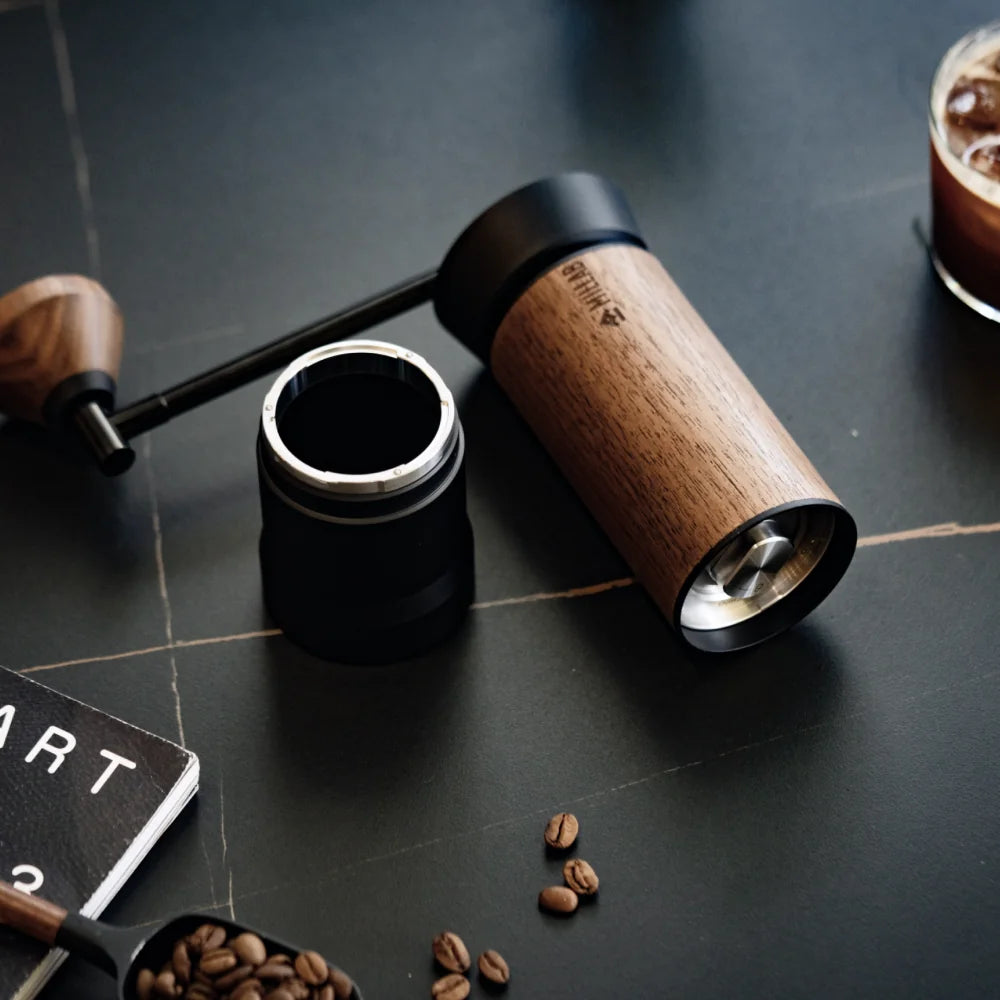 The black and walnut M01 lying on a dark stone counter with its threadless catch cup removed and the crank handle beside it, coffee beans scattered around